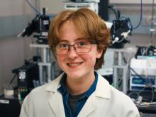 Photo of Will Gaston. He is White with red-brown hair, freckles, and blue eyes. He wears glasses and a lab coat. Behind him, there is scientific equipment.