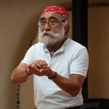 Picture of Laurencio Lopez Nunez storytelling with his hands cupped together in front of him, wearing a red bandana on his head, glasses with a beard and white shirt