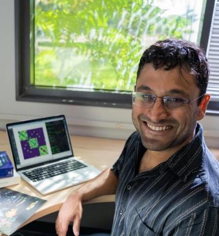 Dr. Rishidev Chaudhuri smiling brightly while sitting at a computer in front a sunny window