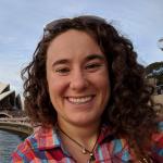A white woman with brown curly hair is smiling outside on a sunny day