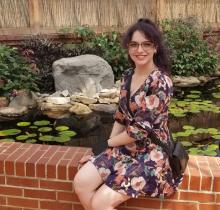 A feminine individual with curly dark hair and a blue floral dress sits on a brick wall in front of a pond full of rocks and lily pads. 